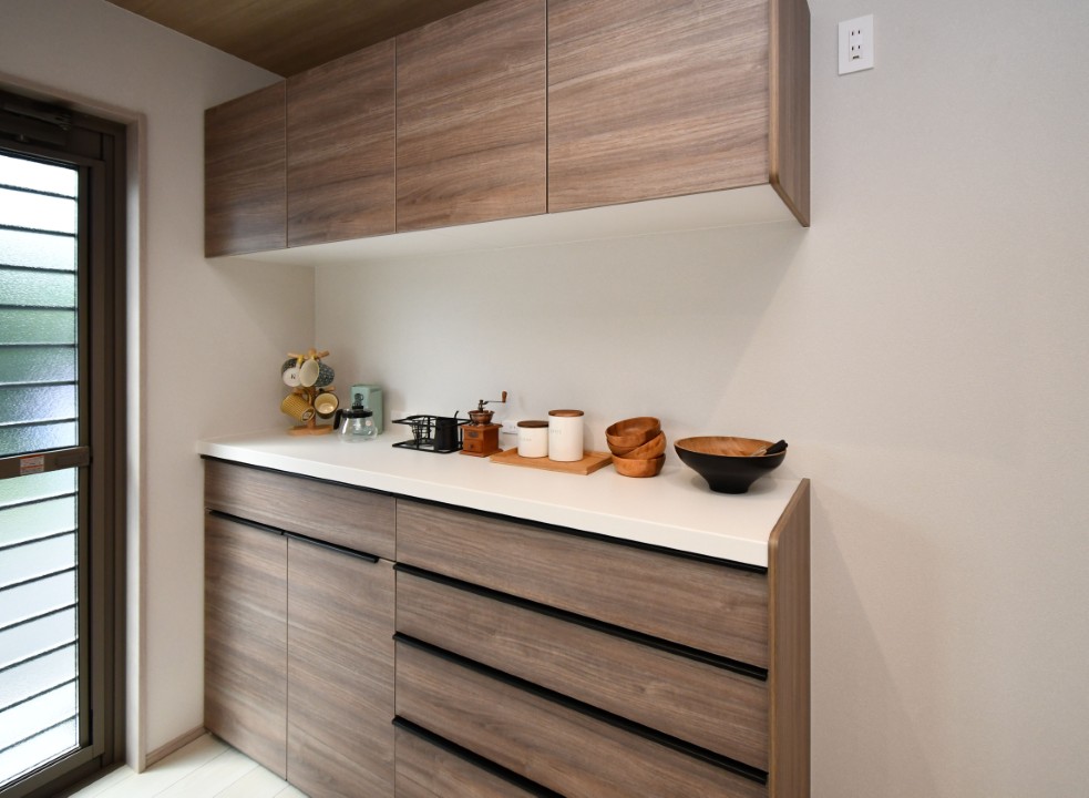 【背面収納にもこだわった美しいキッチン空間】 背面収納には、折下げ天井とトーンを合わせた木目柄を採用し、空間全体に一体感と上質なあたたかみを添えています。
